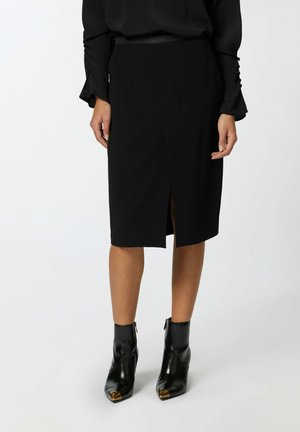 Jupe crayon noire en tissu lisse, dotée d'une fente à l'avant. Associée à des bottines noire avec des accents dorés.