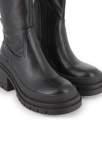 Botas de cuero negras hasta la rodilla con suela de plataforma gruesa, punta redondeada y costuras decorativas. Cuentan con una cremallera lateral para facilitar su uso.