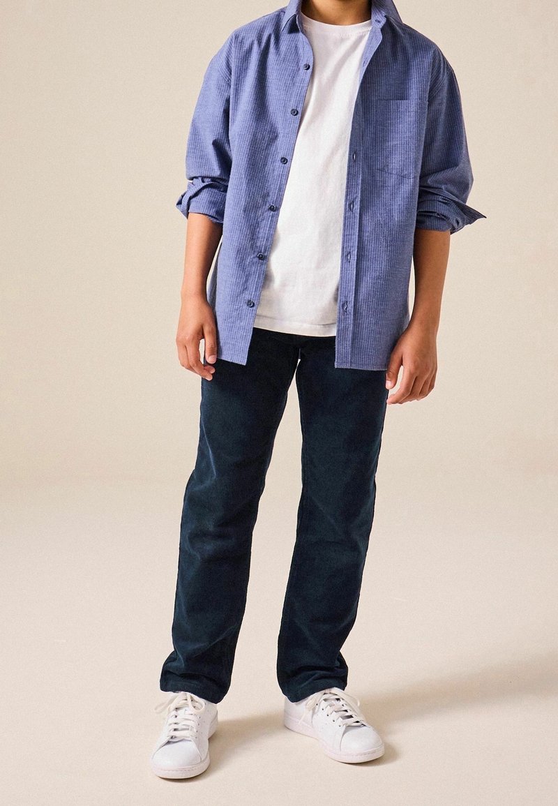 Garçon portant une chemise bleue à boutons par-dessus un t-shirt blanc, un pantalon foncé et des baskets blanches, debout devant un fond clair uni.