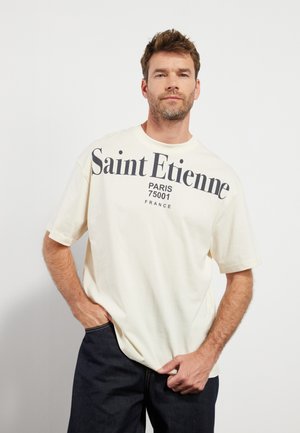 Cremefarvet bomulds T-shirt med korte ærmer, prydet med fed sort tekst "Saint Etienne PARIS 75001 FRANKRIG," afslappet pasform og rund halsudskæring.
