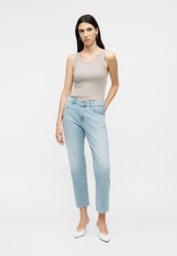 Kvinde står iført en beige ærmeløs top, lyseblå højtaljede jeans, hvide spidse sko og guldringe som øreringe.