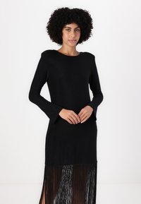 Sort kjole med lange ærmer, rund halsudskæring og frynset kant. Stoffet har en subtil glans, der skaber en elegant silhuet.