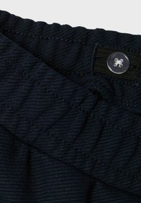 Mörkblått tyg med en texturerad, quiltad mönstring; har en elastisk midja och en klar knapp för knäppning.