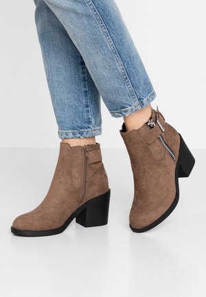 Brune ruskinds støvler med blokhæle, sidelukning med lynlås, spædedetalje, båret med lyseblå op rullede jeans mod en ensfarvet baggrund.