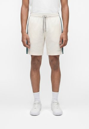 Άνδρας που φοράει μπεζ δίχτυ σορτς με γκρι κορδόνι και σκούρες πράσινες πλευρικές ρίγες, συνδυασμένα με λευκές κάλτσες και λευκά αθλητικά παπούτσια.