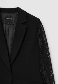 Blazer nero con maniche in pizzo, dotato di chiusura a singolo bottone, rever a punta e tasche frontali. Tessuto liscio con texture in pizzo intricato.