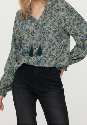 Nainen pukeutuneena löysään, pitkähihaiseen kukkakuosiseen puseroon, jossa on tupsuja, ja korkeavyötäröisiin mustiin farkkuihin, poseeraamassa yhdellä kädellä lantiolla.