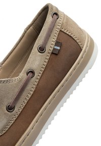 Béžové a hnědé semišové mokasíny s koženým šněrováním, dekorativním prošíváním a bílou texturovanou podrážkou na bílém pozadí.