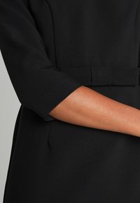 Robe noire avec un corsage ajusté, des manches larges et coupées, et une taille ceinturée. Le tissu semble lisse avec une légère texture.