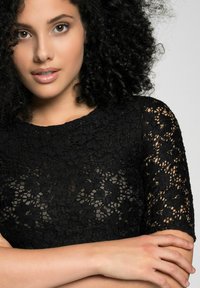 Schwarzes Spitzenoberteil mit kurzen Ärmeln, rundem Ausschnitt und filigranen Blumenmustern. Der Stoff hat ein texturiertes, durchsichtiges Aussehen.