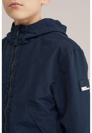 Gyermek sötétkék kapucnis dzsekiben, cipzárral és "Young Originals" felirattal az ujjon, mellkattól felfelé látható.