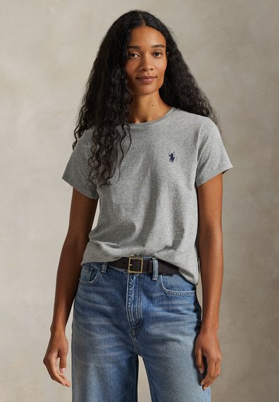 Kvinna med långt lockigt hår som bär en grå t-shirt med logga instoppad i blå jeans med svart bälte, stående mot en enfärgad bakgrund.