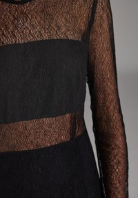 Schwarzes Spitzenoberteil mit transparentem Design und einem undurchsichtigen schwarzen Stoffteil an der Taille. Lange Ärmel und strukturierte Muster sind zu sehen.