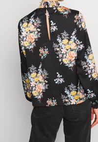 Blouse noire à fleurs avec des motifs colorés, décolleté froncé et une petite ouverture en forme de clé dans le dos. Tissu doux et lisse avec des manches longues.