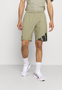 Miestne ľahké olivové šortky s čiernymi akcentovými pruhmi, elastický pás a bočné vrecká; v kombinácii s bielymi športovými topánkami a ponožkami.