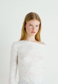 Langarmet, off-shoulder topp i gjennomsiktig hvit stoff med blomsterbroderier og subtil rynking langs siden, glatt tekstur.