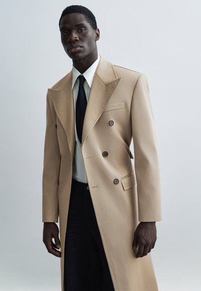 Manteau beige à double boutonnage avec revers à pointe, tissu texturé, grands boutons et coupe ajustée, porté avec une chemise blanche et une cravate noire.