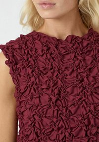 Top bordeaux con un design testurizzato e arricciato. Il tessuto presenta un motivo ondeggiante e un colletto rotondo. Materiale morbido e leggero.