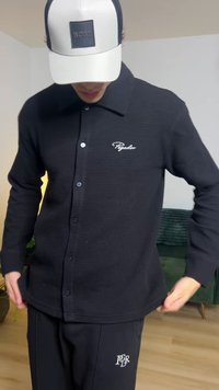 Musta nappipaita, jossa on teksturoiduilla kuvioilla varustettu, terävä kaulus ja vasemmassa rinnassa kirjailtu logo, yhdistettynä tummiin housuihin.