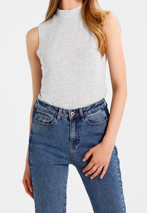 Hihaton vaaleanharmaa korkeakauluksinen paita, yhdistetty korkeavyötäröisiin sinisiin farkkuihin, nainen seisoo yksinkertaisen taustan edessä.