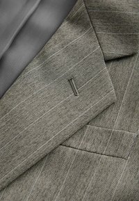 Gros plan sur le revers d'une veste de costume gris rayé et un tissu texturé montrant les détails de la boutonnière et de la poche poitrine.