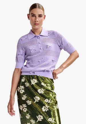 Donna che indossa un polo lavorato a maglia lavanda con dettagli a fiocco e una gonna floreale verde con paillettes, in posa con una mano sul fianco.