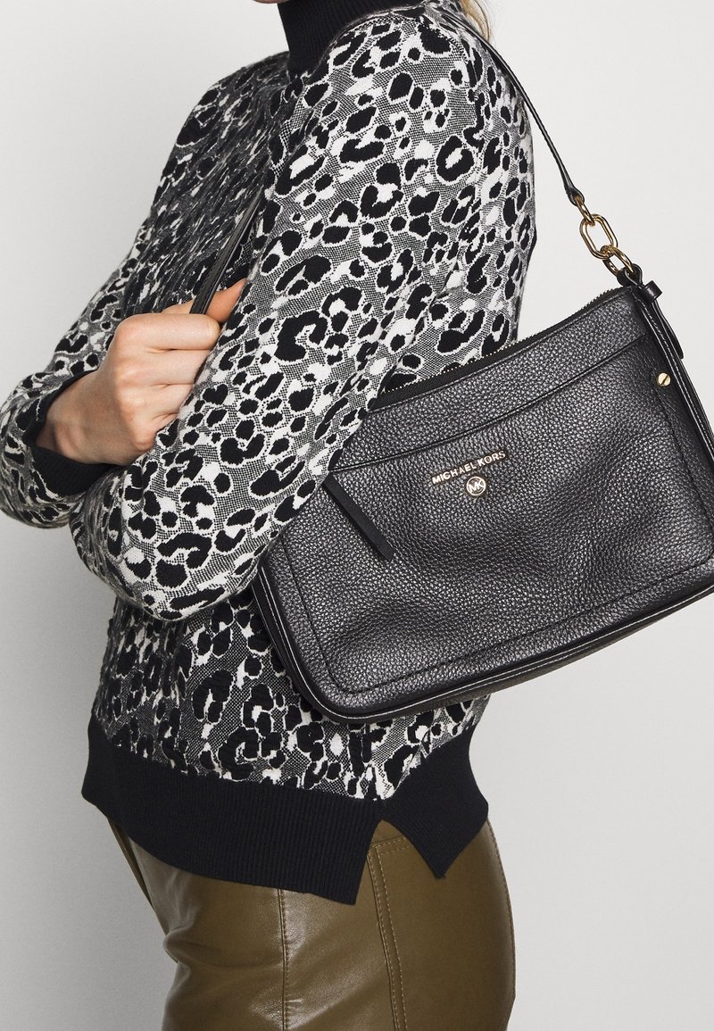 Sort pebret læder håndtaske med lynlåslukning, guldfarvet hardware og flad design, holdt mod en mønstret sweater.