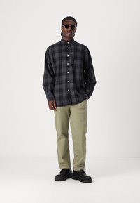 Camisa de cuadros en negro y gris, con tela ligera, combinada con pantalones cargo en verde claro y zapatos negros. Gafas de sol redondas acentúan el look.