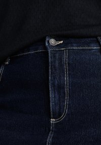 Jeans in denim blu scuro con cuciture visibili e un dettaglio in metallo sul cinturino. Il tessuto ha una consistenza liscia.