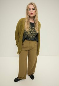 Olivgrüner Strickcardigan mit Fransen, schwarzes Grafik-T-Shirt mit Sonnendesign, hellbraune weite Hosen und schwarze Schuhe.