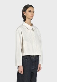 Giacca bianca corta con colletto a punta, maniche lunghe e tasche laterali, abbinata a jeans in denim scuro, esposta su uno sfondo neutro.