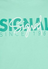 Camiseta verde menta con gran texto blanco "SIGNAL", que presenta una combinación de tipografía en negrita y cursiva, y una etiqueta "DESDE 1966" debajo.