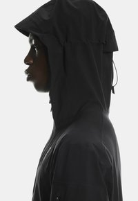 Černá kapucová bunda s elegantním designem. Obsahuje strukturovanou tkaninu, elastickou šňůrku na kapuci a detaily se zipovými postranními kapsami.