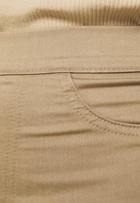 Beige denimová látka s detailním záběrem na kapsu, viditelným šitím a hladkou texturou, s ribbovaným pasem nahoře.