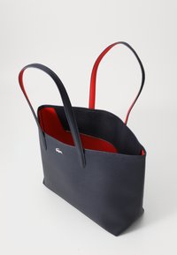 Sac cabas en cuir bleu marine avec une texture lisse, un intérieur rouge, de longues poignées et un accent logoté argenté sur le côté. Design à ouverture supérieure.