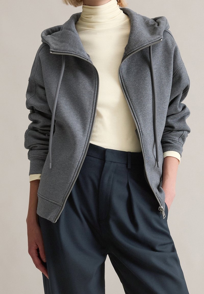 Personne portant un sweat à capuche gris zippé sur un col roulé crème et un pantalon large bleu marine foncé, une main dans la poche.