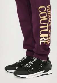 Sapatos de desporto pretos com acentos e logotipo brancos, apresentando uma parte superior texturizada, atacadores elásticos e uma sola robusta. Usados com calças de treino roxas.
