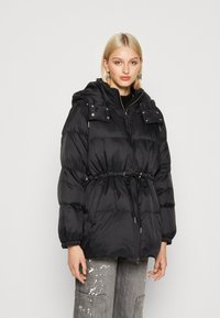 Veste matelassée noire avec une taille cintrée, fermeture éclair et capuche amovible. Équipée de poignets élastiques et de deux poches poitrine avec boutons-pression.