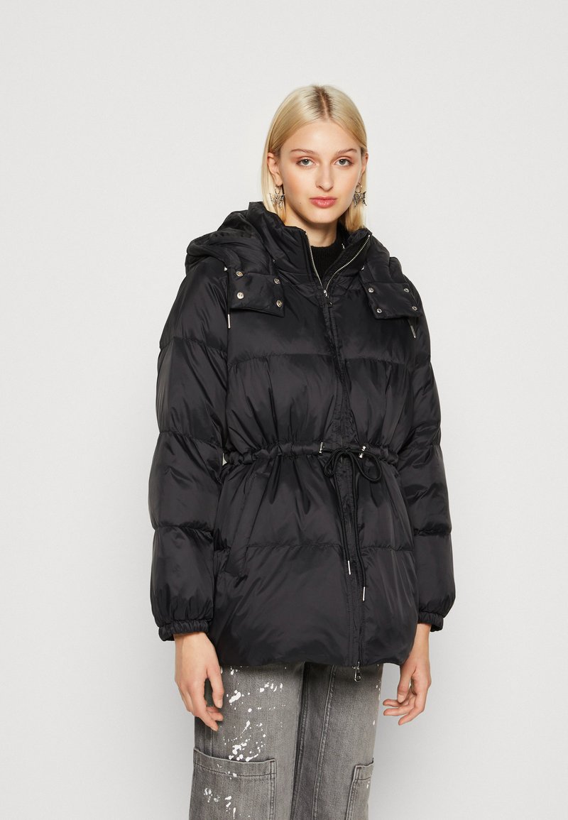 Veste matelassée noire avec une taille cintrée, fermeture éclair et capuche amovible. Équipée de poignets élastiques et de deux poches poitrine avec boutons-pression.