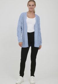 Svijetloplava pletena cardigan jakna labavog kroja, s rebrastim rubovima i dekorativnim uzorcima. Nosena preko bijele bluze i uparena s crnim hlačama.