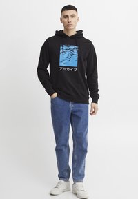 Felpa nera con un design grafico blu che presenta il testo giapponese "アーカイブ". Indossata con jeans in denim azzurro chiaro e sneaker bianche.