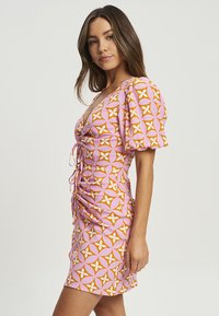 Korte, getailleerde jurk van roze stof met een oranje en witte geometrische print. Heeft pofmouwen en een veterdetail aan de voorkant.