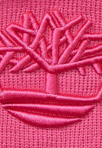 Rosa broderad logotyp med en stiliserad trädt som har grenar och rötter, placerad på en texturerad rosa tygbakgrund.