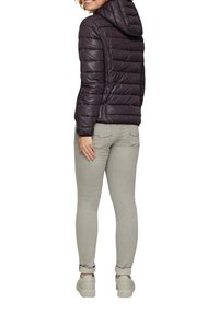 Doudoune violette foncée avec capuche, présentant un design matelassé et une légère brillance ; associée à un jean gris et des chaussures claires.