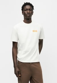 Άνδρας που στέκεται, φοράει λευκό t-shirt Dickies και καφέ παντελόνι μπροστά σε απλό φωτεινό φόντο, τα χέρια χαλαρά στα πλάγια.