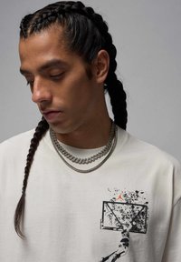 Cremefarbenes T-Shirt mit einem Basketballgrafik auf der linken Brustseite, das eine schwarze Silhouette zeigt. Das Modell trägt zwei silberne Ketten und hat geflochtene Haare.