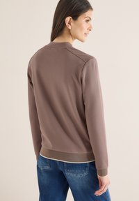 Femme vue de dos portant un pull taupe à manches longues avec poignets et ourlet côtelés, associé à un jean en denim bleu sur un fond uni.