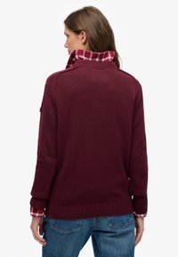 Woman facing away wearing a dark red knit sweater over a red and white plaid shirt, paired with blue denim jeans against a plain white background.