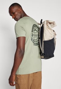 Lys grøn t-skjorte med et rundt logo på baksiden, kombinert med en beige og svart ryggsekk med teksturert design og justerbare stropper.