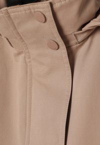 Gros plan sur le col d'un manteau beige avec deux boutons-pression assortis et des coutures visibles le long des bords des coutures.